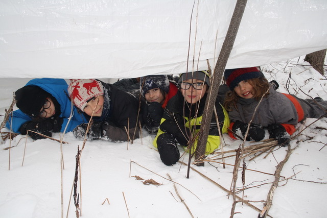 5 garçons de l'unité des Aigles, couchés dans la neige sous une bâche, démontrant comment se protéger des intempéries, en hiver 2020