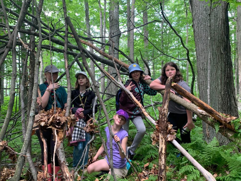 Cinq jeunes filles dans la forêt, derrière un abri de divers morceaux de bois.
