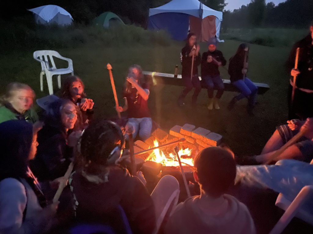 Des enfants autour d'un feu de camp, en soirée. Les jeunes ont des bâtons et font griller des guimauves.